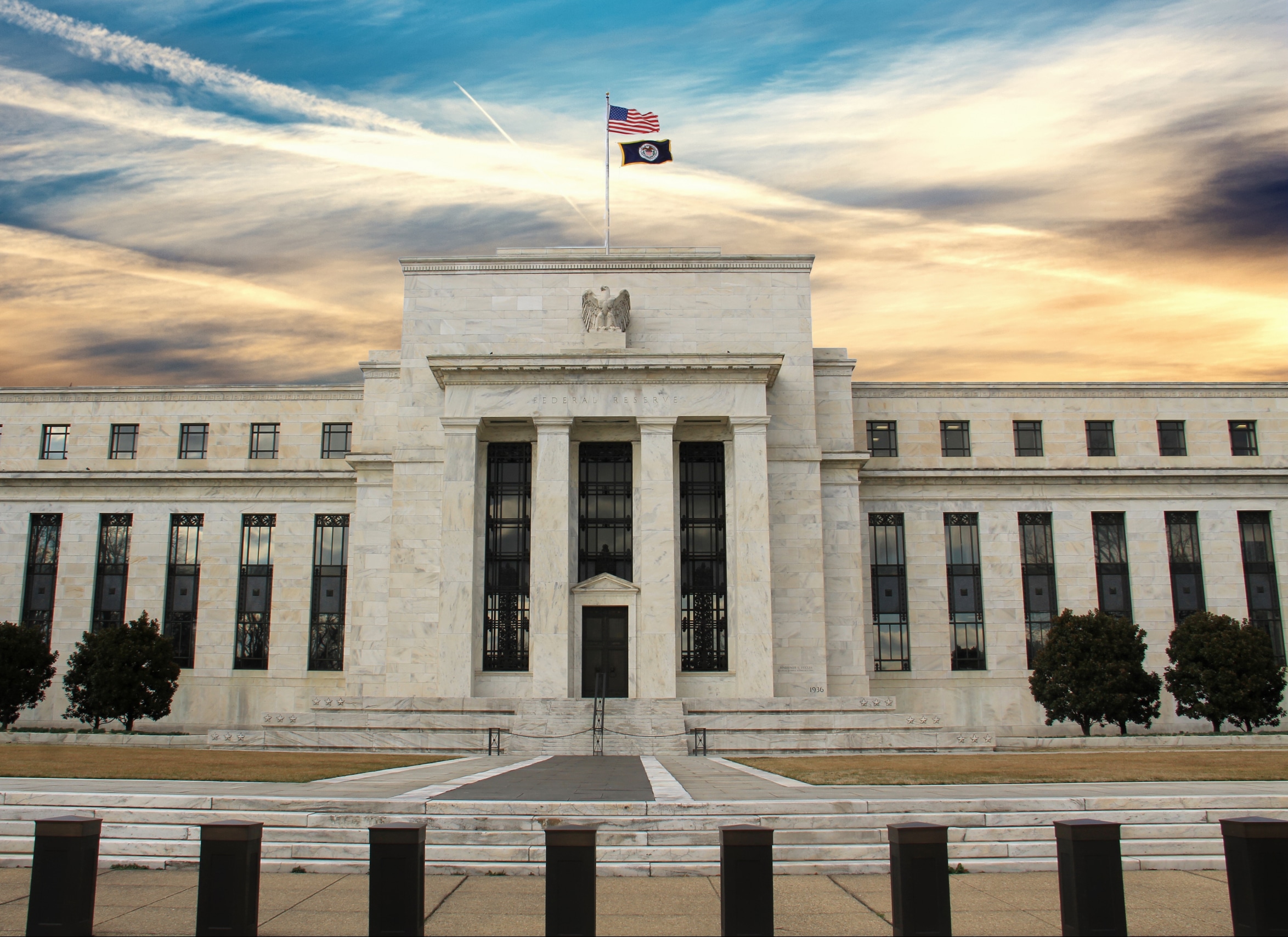 The image shows the exterior of the Federal Reserve Building, a large rectangular structure with a central entrance, tall columns, and an American flag atop. The sky is partly cloudy with blue, orange, and yellow hues.