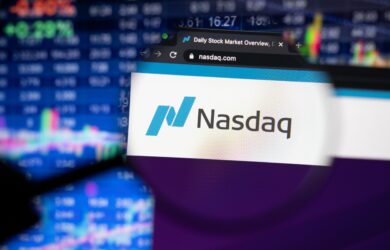 Close-up of a computer screen displaying the Nasdaq website with its logo in focus through a magnifying glass. In the background, blurred stock market graphs and charts are visible.