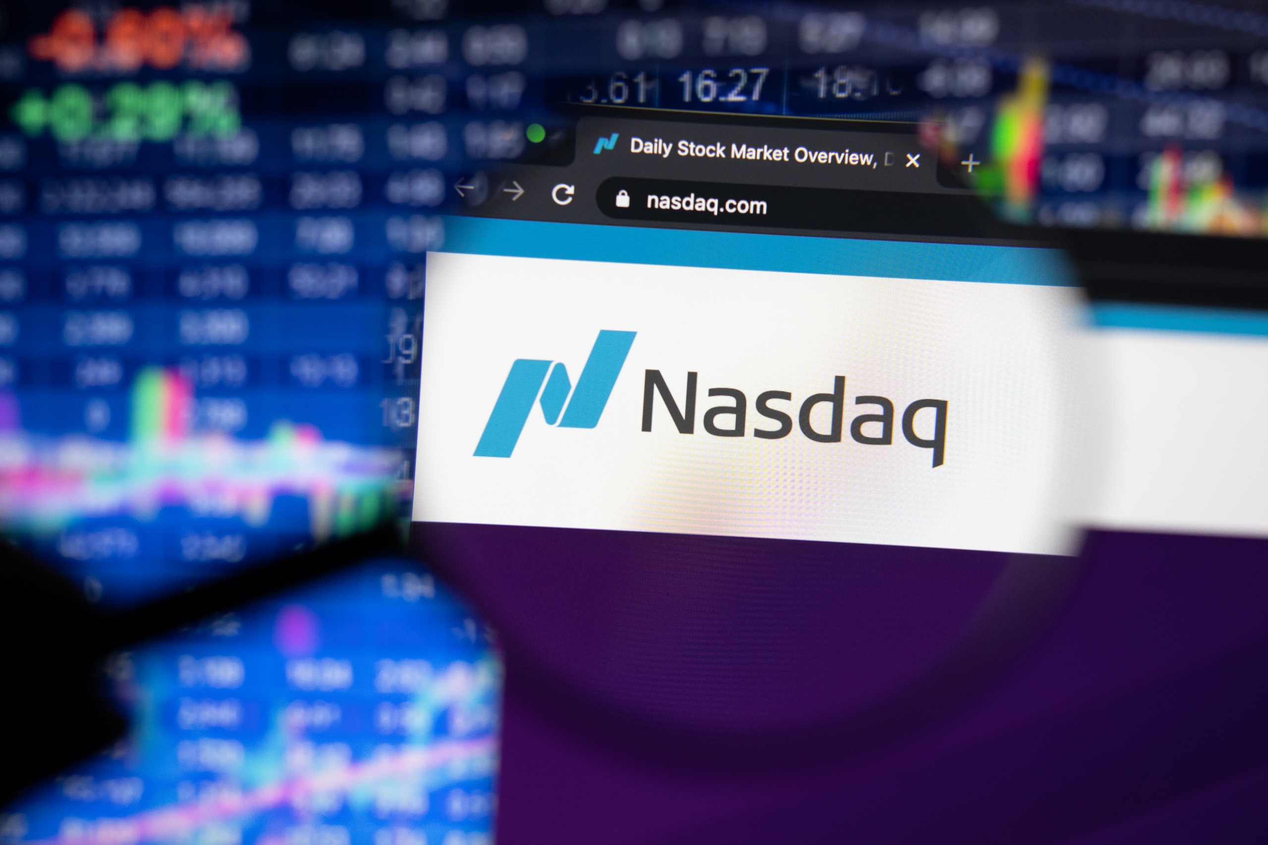 Close-up of a computer screen displaying the Nasdaq website with its logo in focus through a magnifying glass. In the background, blurred stock market graphs and charts are visible.