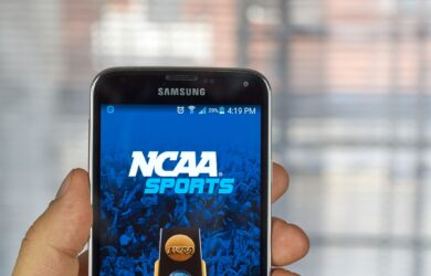 A person holds a Samsung smartphone displaying the NCAA Sports app screen. The background shows a blurred window with natural light.