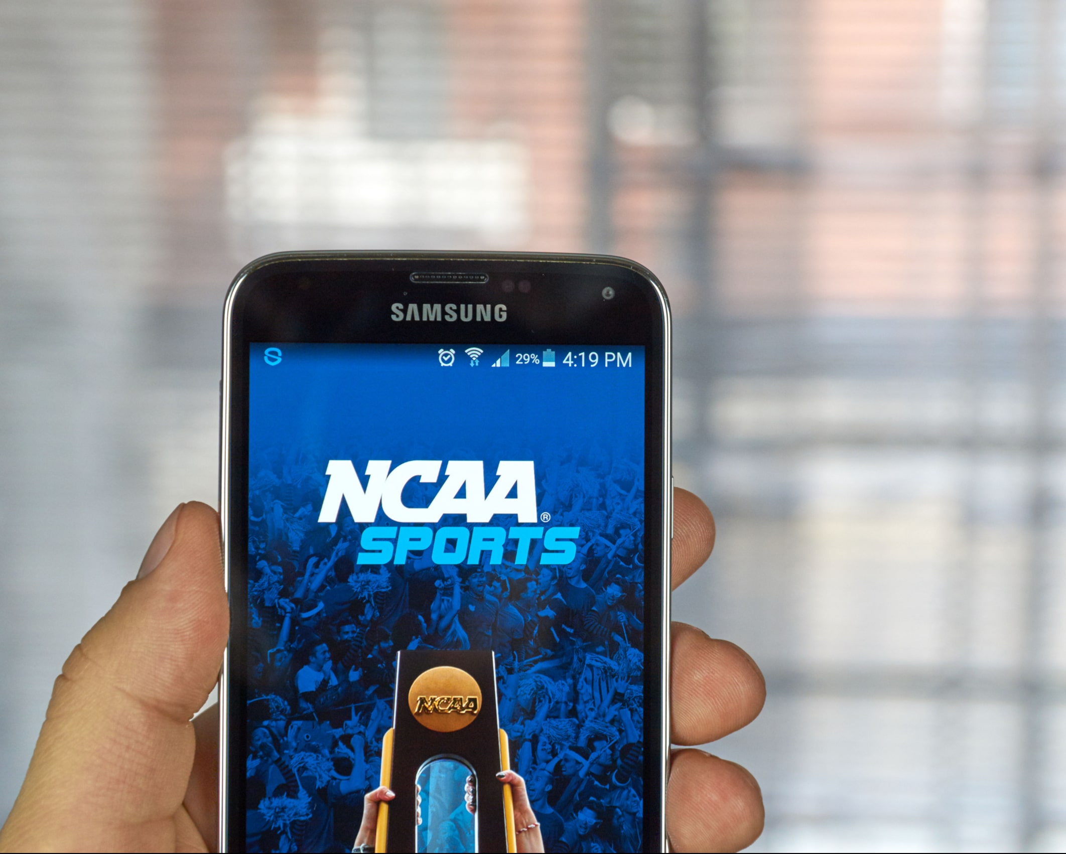 A person holds a Samsung smartphone displaying the NCAA Sports app screen. The background shows a blurred window with natural light.