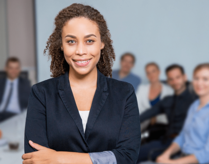 A confident person with curly hair and a navy blazer stands smiling with arms crossed. In the background, a group of people is seated at a table, engaging in a meeting. The setting appears professional and collaborative.