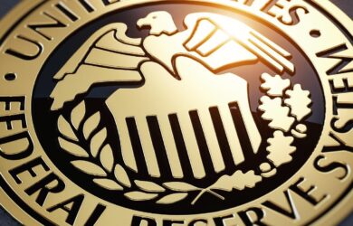 Gold emblem of the United States Federal Reserve System featuring an eagle, shield, and olive branches, with light reflecting on its surface. Engraved text encircles the emblem.