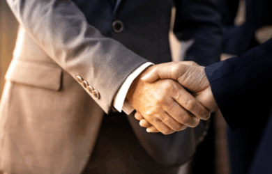 Two individuals in business attire are shaking hands. The person on the left is wearing a gray suit, and the person on the right is wearing a dark suit. The background is softly blurred, emphasizing the handshake as a symbol of agreement or partnership.