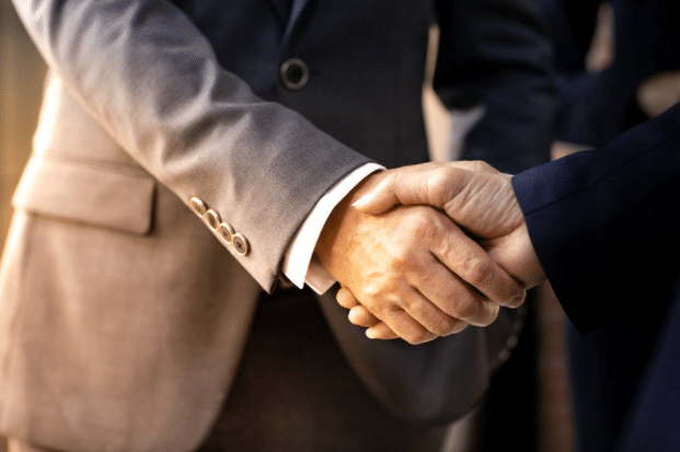 Two individuals in business attire are shaking hands. The person on the left is wearing a gray suit, and the person on the right is wearing a dark suit. The background is softly blurred, emphasizing the handshake as a symbol of agreement or partnership.