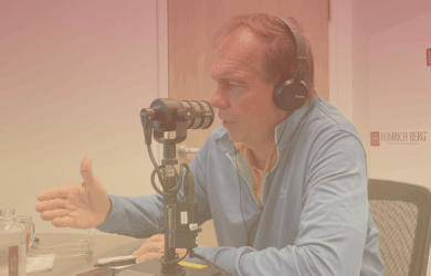 A man in headphones speaks into a microphone in a podcast studio. He gestures with his hand while seated at a table with a water bottle, notepad, and company branding on the wall behind him.