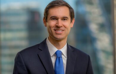 A man in a suit and blue tie is smiling, standing in front of a blurred cityscape background.