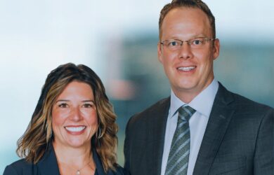 A woman and man are smiling at the camera. The woman has shoulder-length hair and is wearing a dark blazer. The man is wearing glasses, a suit, and a tie. The background is blurred and light-colored.