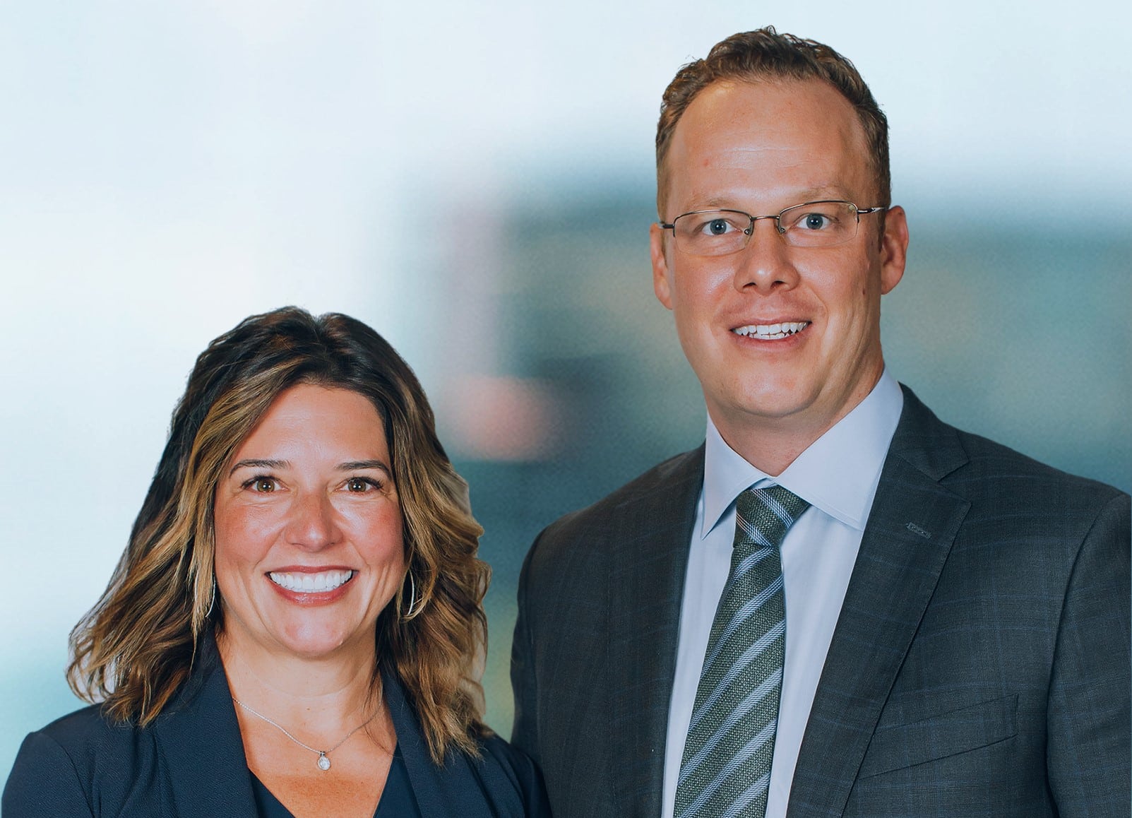 A woman and man are smiling at the camera. The woman has shoulder-length hair and is wearing a dark blazer. The man is wearing glasses, a suit, and a tie. The background is blurred and light-colored.