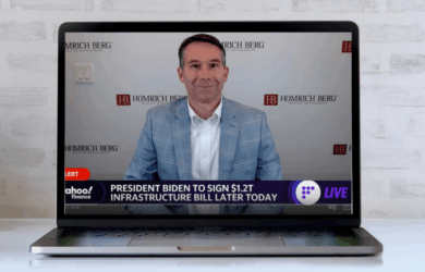 A man in a light blue suit sits in front of a laptop, with a virtual background displaying the Homrich Berg logo. The screen shows a news ticker about President Biden signing a $1.2T infrastructure bill later today.