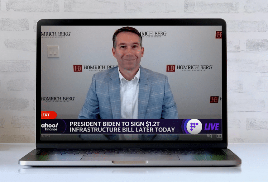 A man in a light blue suit sits in front of a laptop, with a virtual background displaying the Homrich Berg logo. The screen shows a news ticker about President Biden signing a $1.2T infrastructure bill later today.