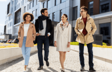 Four people walk on a paved path beside modern buildings. They wear coats and scarves, suggesting cool weather. The group appears engaged in conversation, enjoying a casual stroll. The background features grassy areas and a partly cloudy sky.