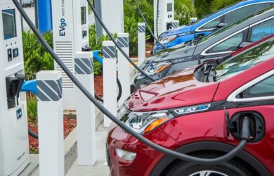 Electric vehicles parked at charging stations, connected to chargers. The scene includes green landscaping and multiple cars in different colors, with charging cables plugged in.