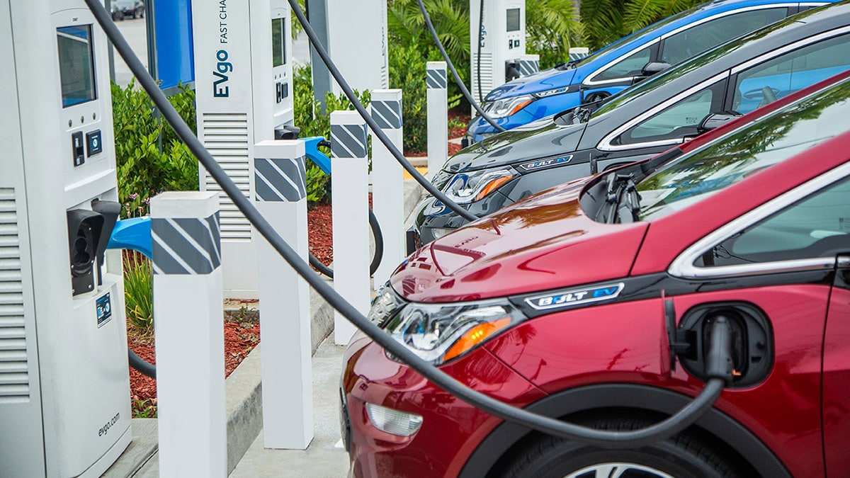 Electric vehicles parked at charging stations, connected to chargers. The scene includes green landscaping and multiple cars in different colors, with charging cables plugged in.