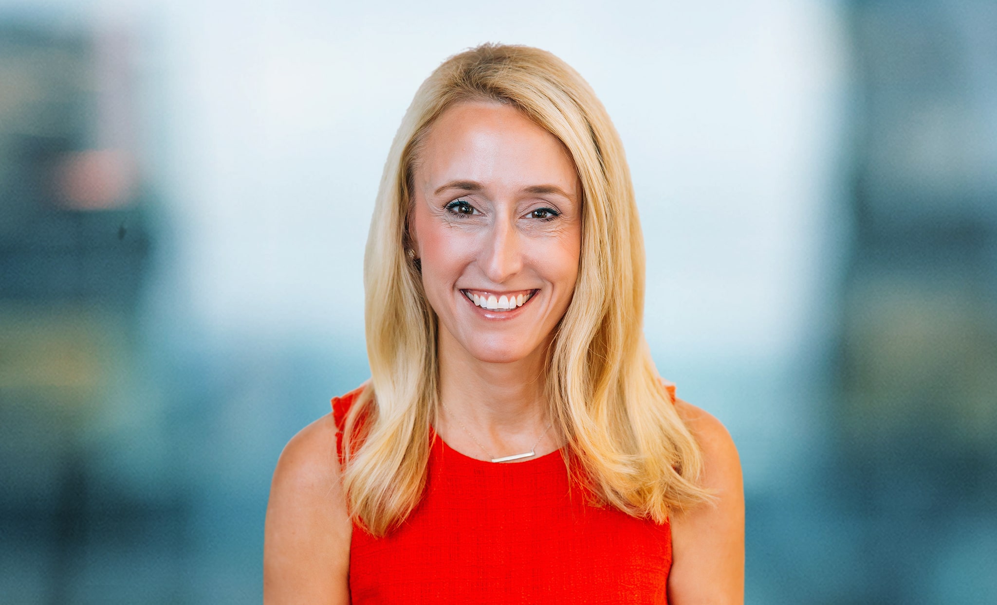 A woman with long blonde hair is smiling, wearing a sleeveless red dress. She is standing against a blurred indoor background.
