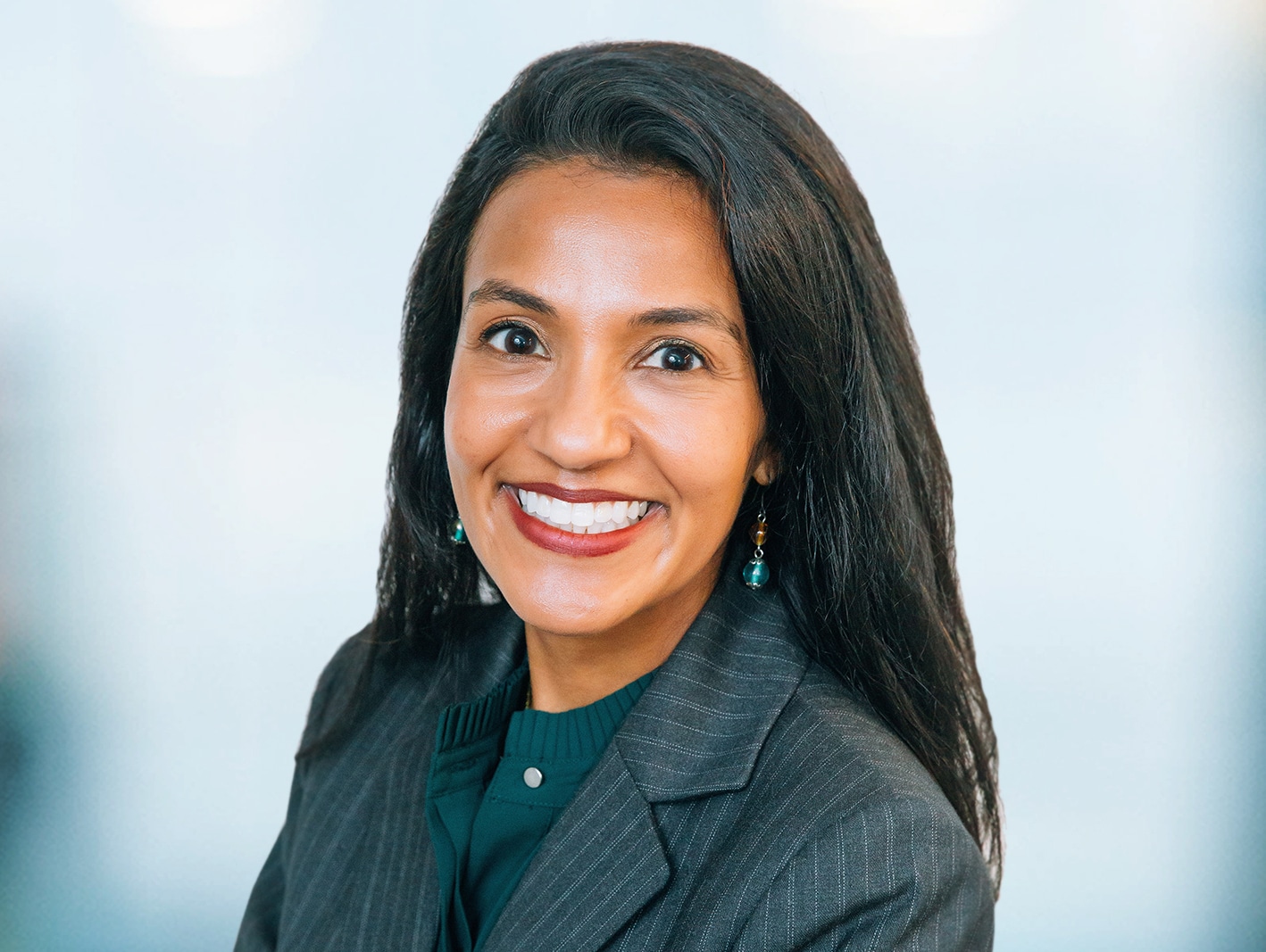 A person with long dark hair, wearing a gray blazer and green top, smiles warmly at the camera. The background is softly blurred, creating a professional and friendly atmosphere.