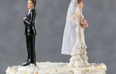 A wedding cake topper featuring a bride and groom figurine standing back to back. The groom is in a black suit, and the bride is in a white dress with a veil, holding a bouquet. The cake is decorated with white icing and pearls.