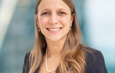 Smiling woman with long hair wearing a dark blazer, red top, and pearl necklace. She is standing against a blurred, light blue background.