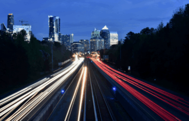 A long-exposure shot of a highway at dusk shows streaks of white and red lights from vehicles in motion. The illuminated city skyline is visible in the background against a deep blue sky. Trees line the side of the road, adding a touch of nature.