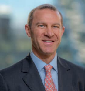 A man in a suit with a light blue shirt and a pink tie smiles at the camera. The background is blurred, showing large windows and hints of a cityscape.