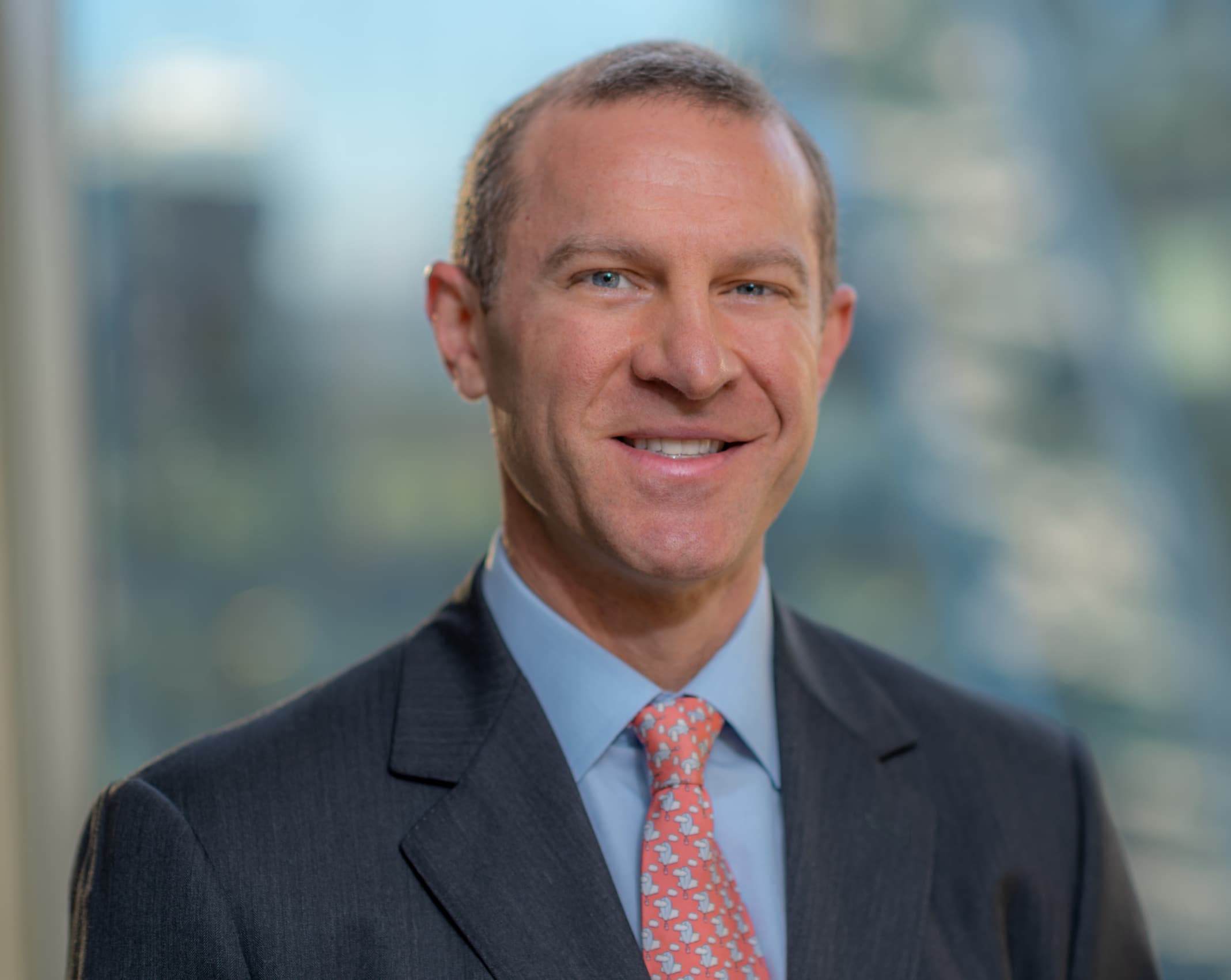 A man in a suit with a light blue shirt and a pink tie smiles at the camera. The background is blurred, showing large windows and hints of a cityscape.