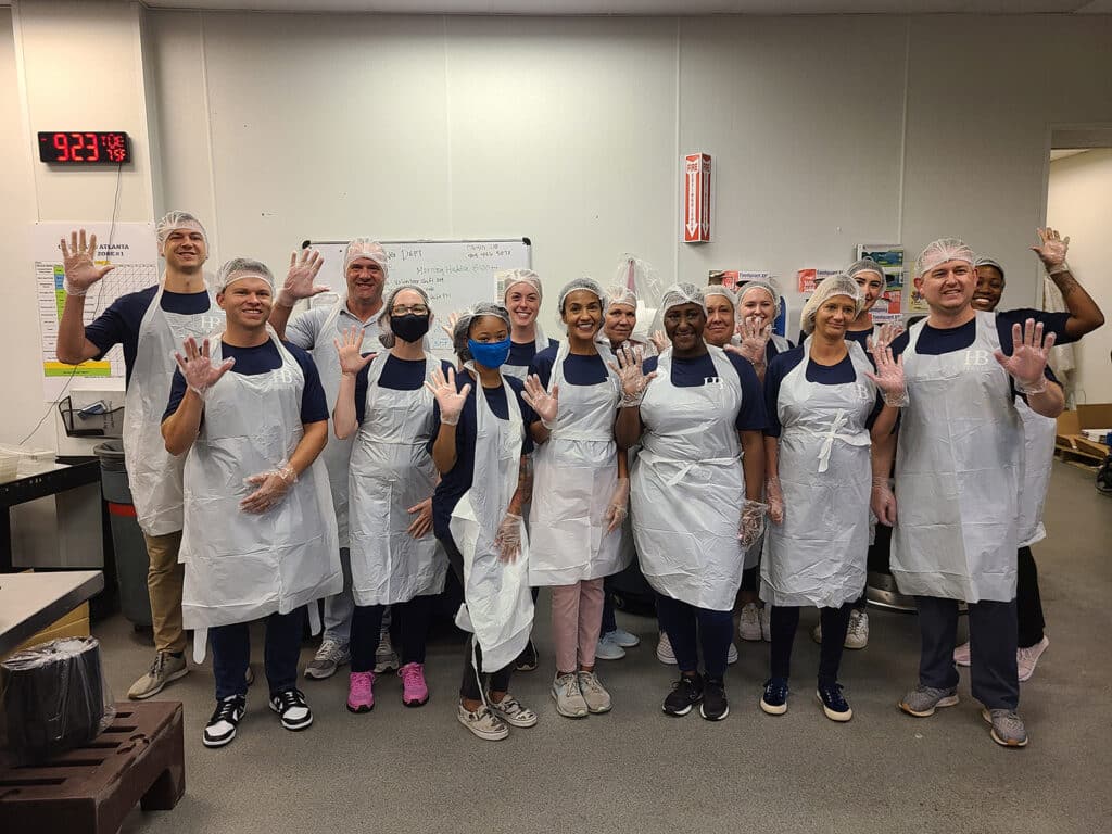 A group of people wearing hairnets, plastic aprons, and gloves stand in a kitchen-like setting, smiling and waving at the camera. A clock on the wall shows 9:23. Whiteboard and kitchen supplies are visible in the background.