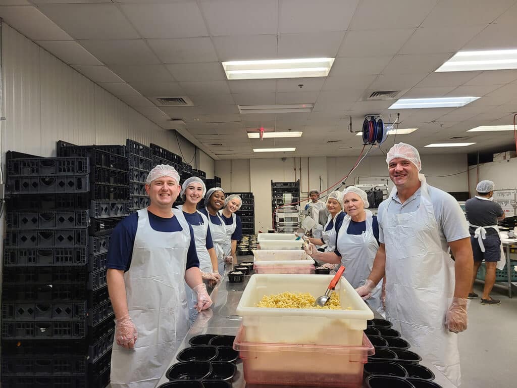 A group of people wearing hairnets and aprons stand in a food preparation area. They are smiling and gathered around tables with trays of food, including pasta. The background shows shelves and crates in the room.