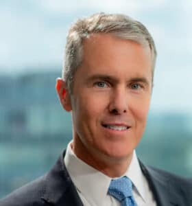 A man with short gray hair wearing a dark suit, light blue shirt, and blue polka dot tie stands in front of a blurred office background.