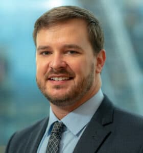 A person with short brown hair and a beard is smiling while wearing a gray suit, a light blue shirt, and a patterned tie. The background is softly blurred but appears to depict a modern interior setting.