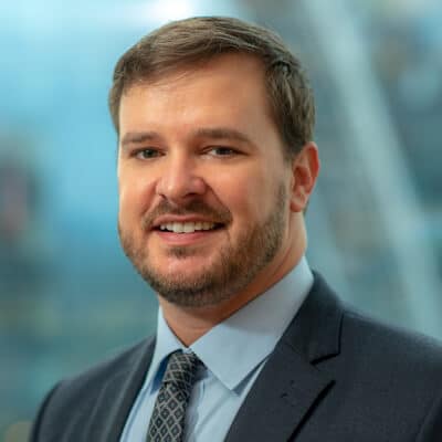 A person with short brown hair and a beard is smiling while wearing a gray suit, a light blue shirt, and a patterned tie. The background is softly blurred but appears to depict a modern interior setting.