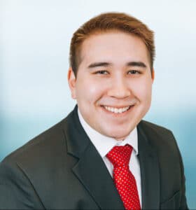 A person in a black suit and red polka dot tie smiles at the camera. The background is a soft blue gradient.