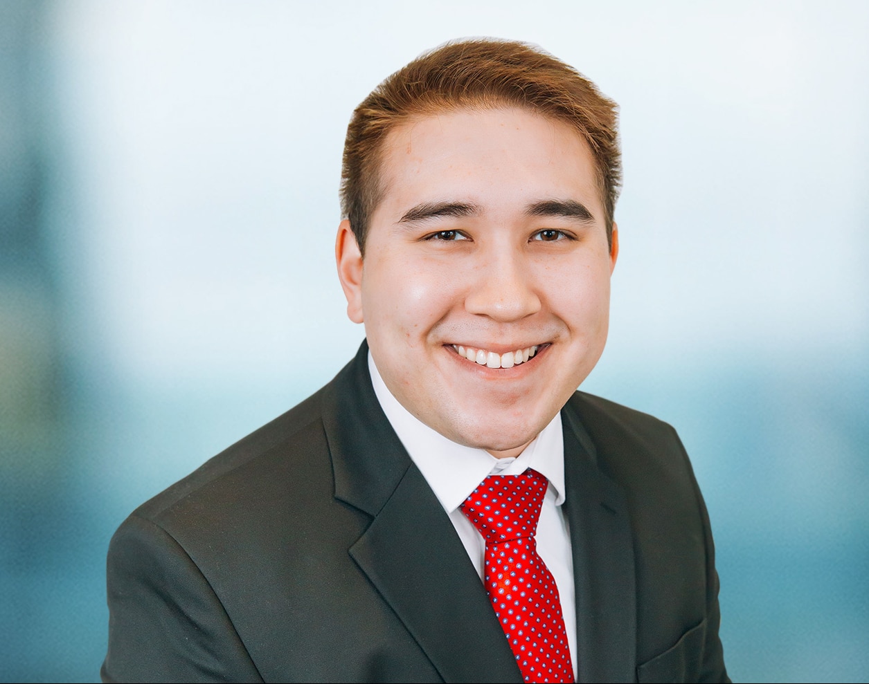 A person in a black suit and red polka dot tie smiles at the camera. The background is a soft blue gradient.