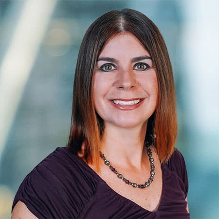 Woman with shoulder-length, straight brown hair, smiling, wearing a dark purple top and a beaded necklace. Background is blurred with hints of blue and yellow, giving a professional, warm atmosphere.