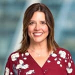 A person with shoulder-length brown hair smiles while wearing a red floral-patterned blouse. The background is blurred, featuring shades of blue and gray.
