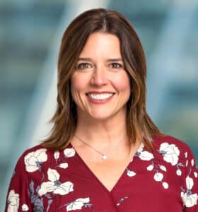 A person with shoulder-length brown hair smiles while wearing a red floral-patterned blouse. The background is blurred, featuring shades of blue and gray.
