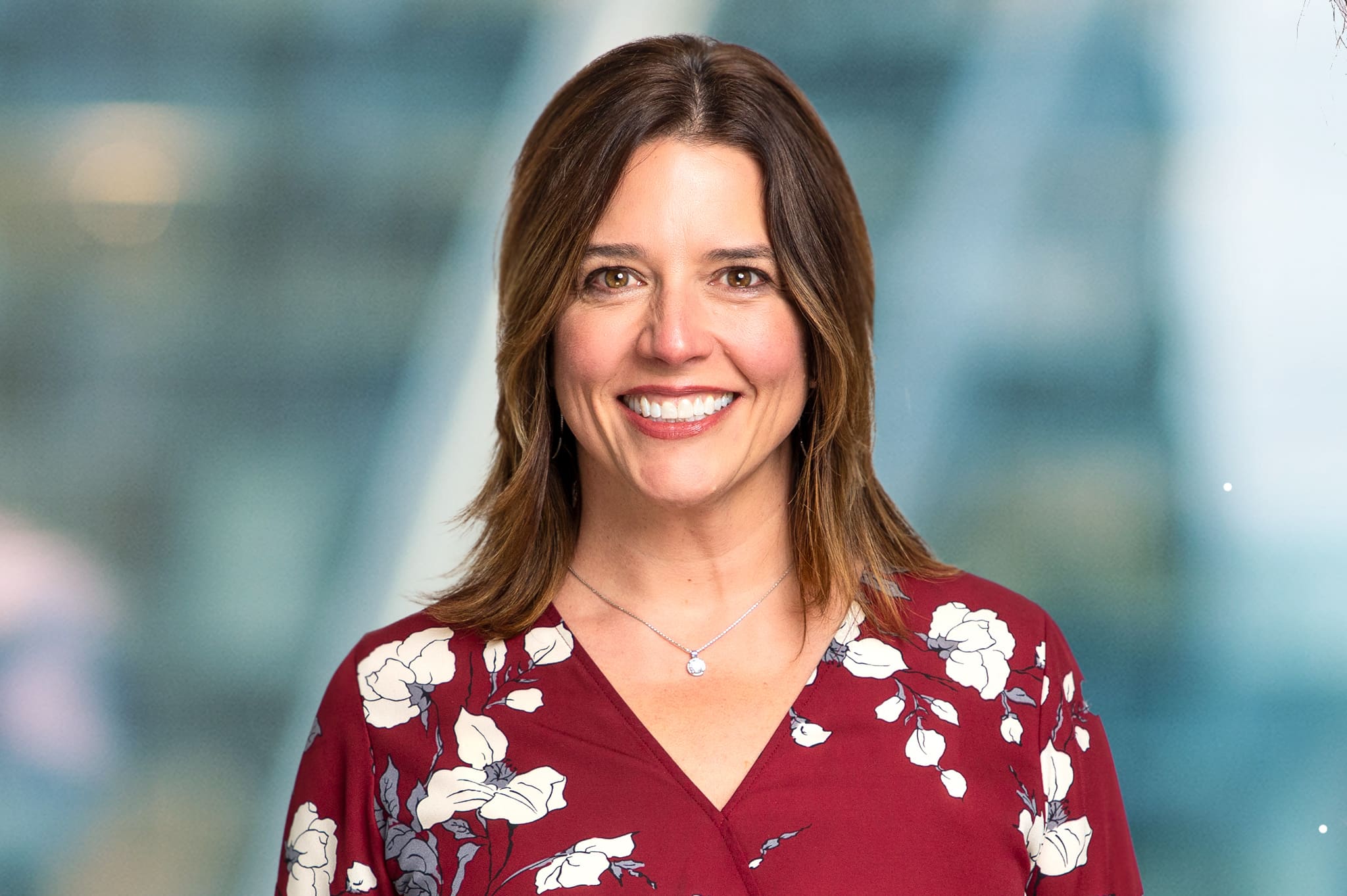 A person with shoulder-length brown hair smiles while wearing a red floral-patterned blouse. The background is blurred, featuring shades of blue and gray.