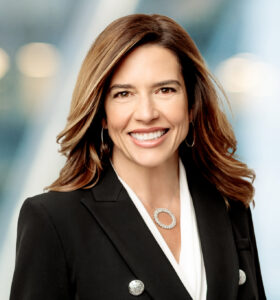 A woman with long brown hair wearing a black blazer and white blouse smiles at the camera. The background is blurred with soft lights and blue tones.
