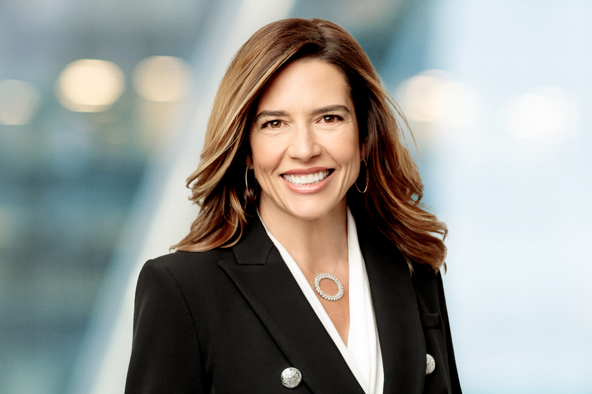 A woman with long brown hair wearing a black blazer and white blouse smiles at the camera. The background is blurred with soft lights and blue tones.