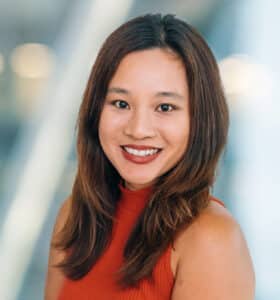 A person with long brown hair smiling, wearing a sleeveless red top. The background is softly blurred with circular lights, creating a light and airy atmosphere.