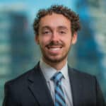 A person with curly hair and a beard is smiling while wearing a dark suit, light shirt, and striped tie. The background is blurred, showing a cityscape with tall buildings.