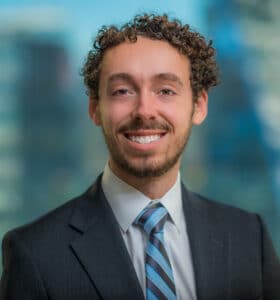 A person with curly hair and a beard is smiling while wearing a dark suit, light shirt, and striped tie. The background is blurred, showing a cityscape with tall buildings.