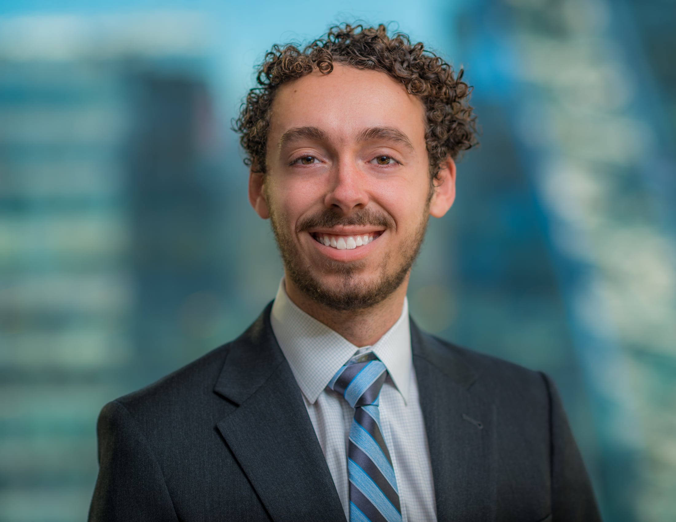 A person with curly hair and a beard is smiling while wearing a dark suit, light shirt, and striped tie. The background is blurred, showing a cityscape with tall buildings.