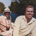 Two men smiling while boating on a narrow waterway. The man on the left wears a cream sweater, white hat, and has binoculars around his neck. The man on the right wears a cream sweater and has blond hair. Lush greenery lines the waterway.