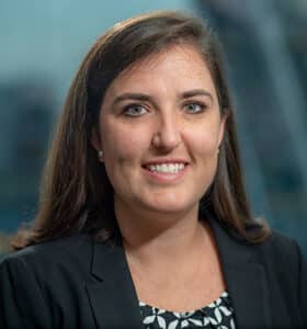 A woman with shoulder-length brown hair smiles at the camera. She is wearing a black blazer over a patterned blouse. The background is softly blurred.