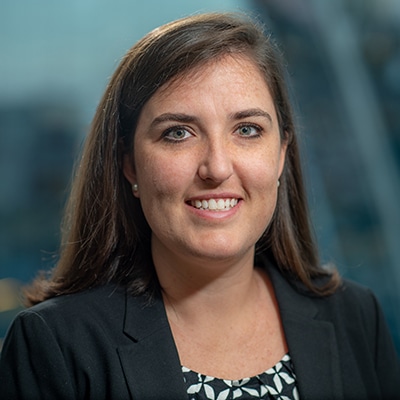 A woman with shoulder-length brown hair smiles at the camera. She is wearing a black blazer over a patterned blouse. The background is softly blurred.