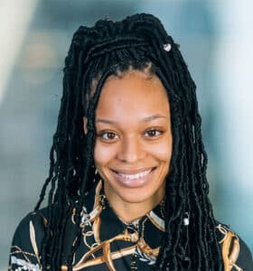 A person with long braided hair is smiling at the camera. They are wearing a patterned shirt against a blurred background.