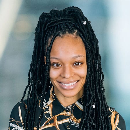 A person with long braided hair is smiling at the camera. They are wearing a patterned shirt against a blurred background.
