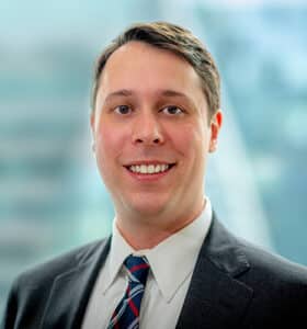 A man in a suit and tie smiles at the camera. The background is blurred, suggesting an office setting.