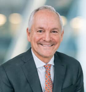 An older man in a suit and patterned tie smiles at the camera. The background is blurred, displaying a neutral-toned indoor setting.