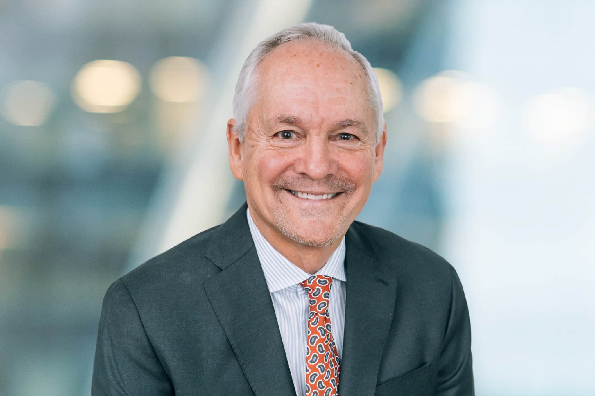 An older man in a suit and patterned tie smiles at the camera. The background is blurred, displaying a neutral-toned indoor setting.